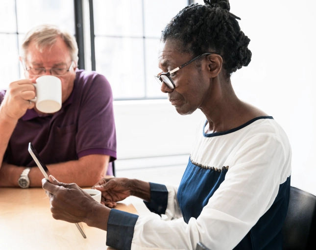 Older couple sitting at a table together. The woman look at a phone screen while the man sips coffee.